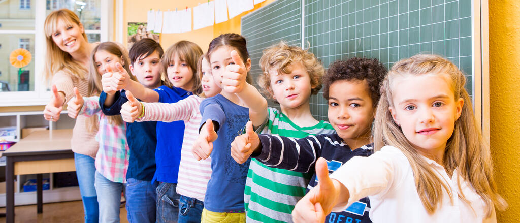 Lehrerin und Grundschulkinder vor der Tafel zeigen Daumen hoch